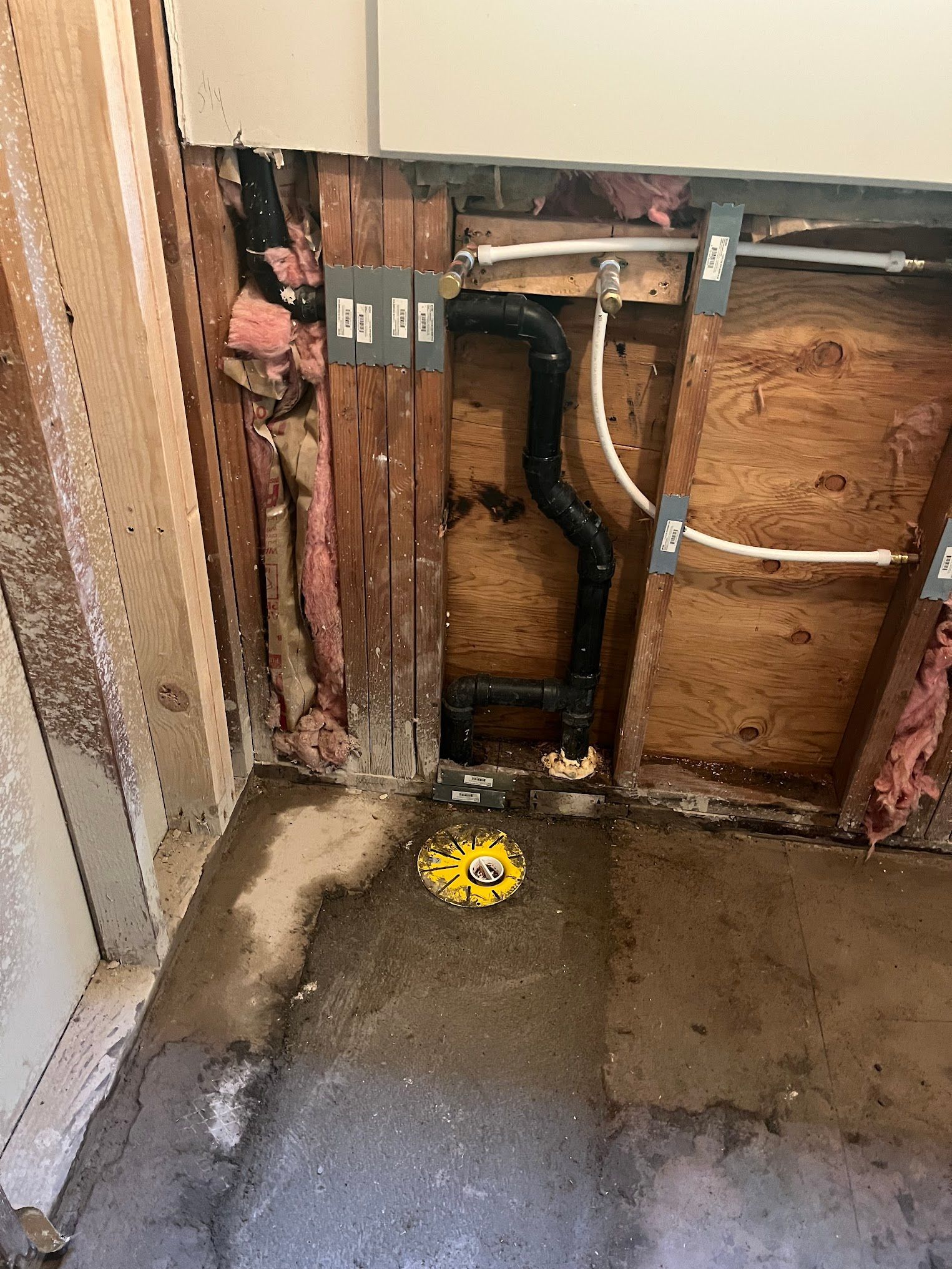 Exposed plumbing in a bathroom with black pipes, white water lines, and a yellow drain cover.