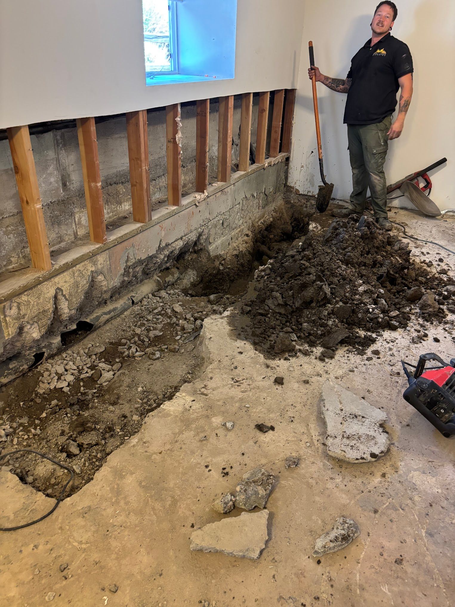 Man digging in a basement. Exposed wall studs above concrete foundation; dirt and debris cover floor.
