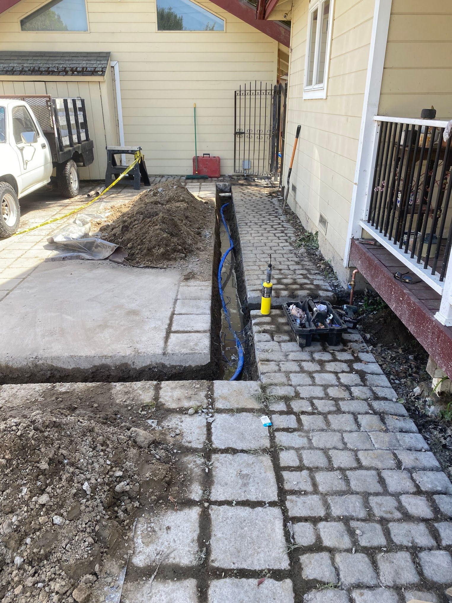 Trench dug along paved walkway. Dirt pile, blue pipes, and tools visible. House and truck in the background.