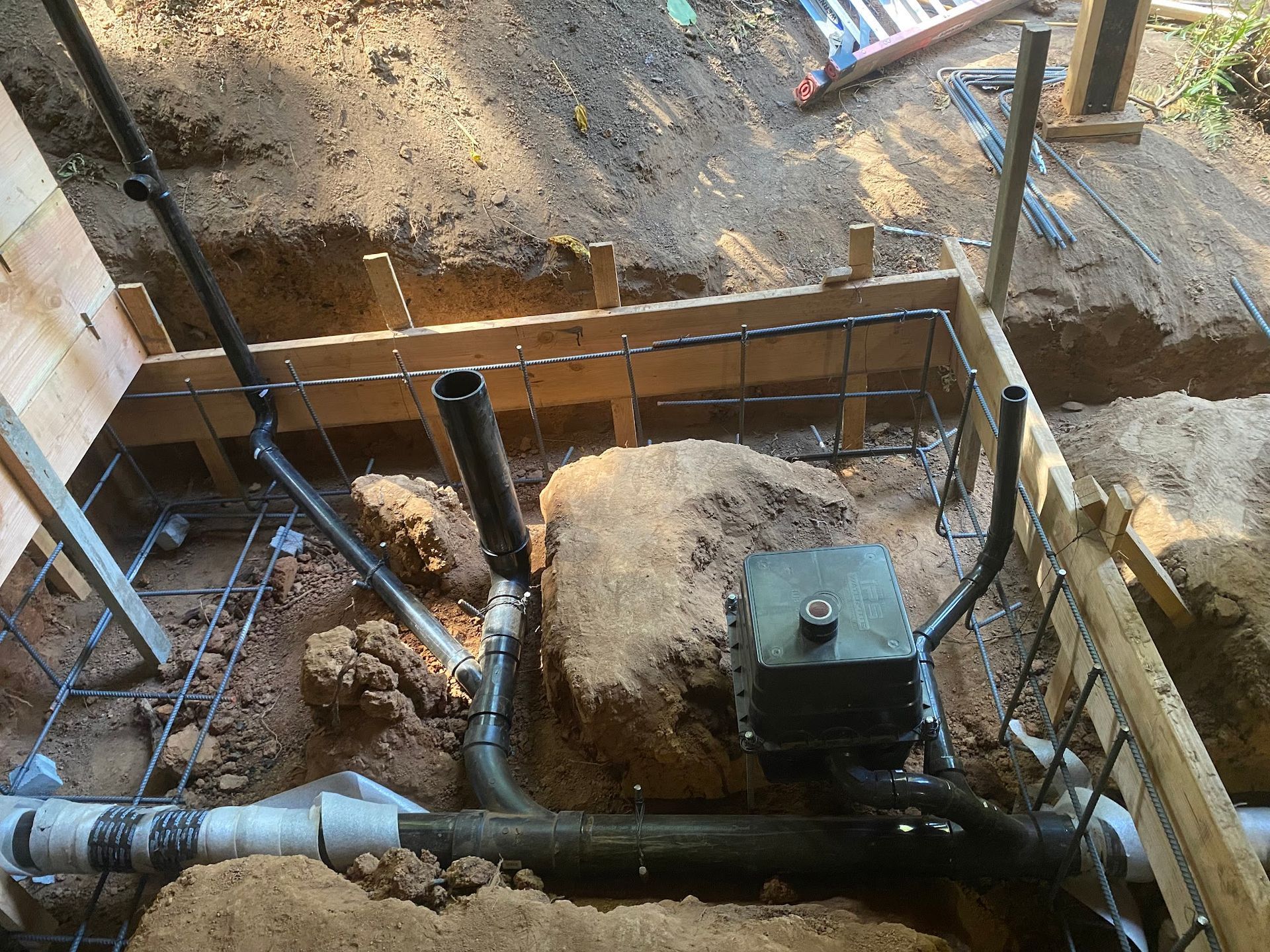 Excavation site with plumbing and electrical work; black pipes, a grey box, and wood framing.