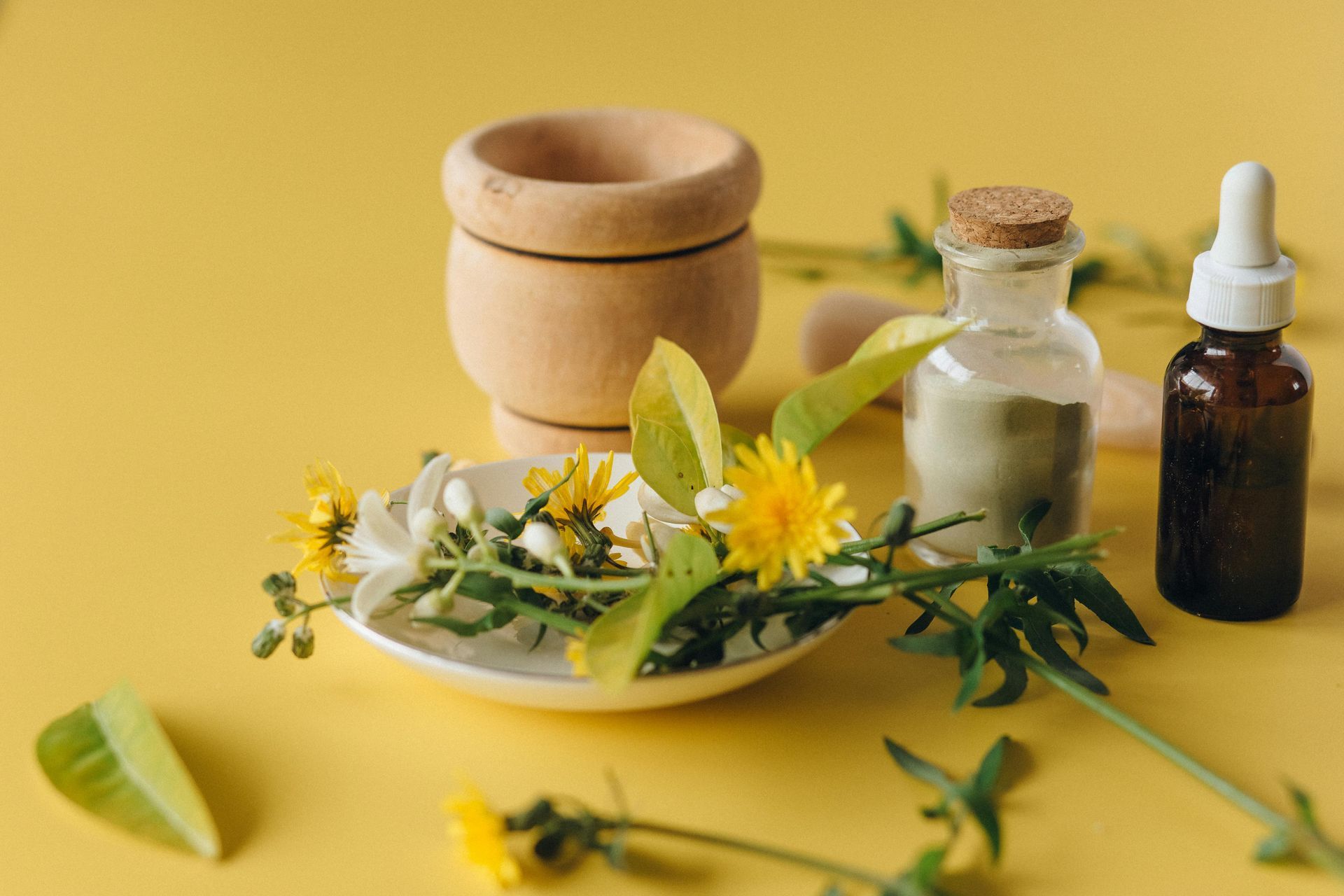 A plate of flowers , a mortar and pestle , a bottle of powder and a dropper on a yellow table