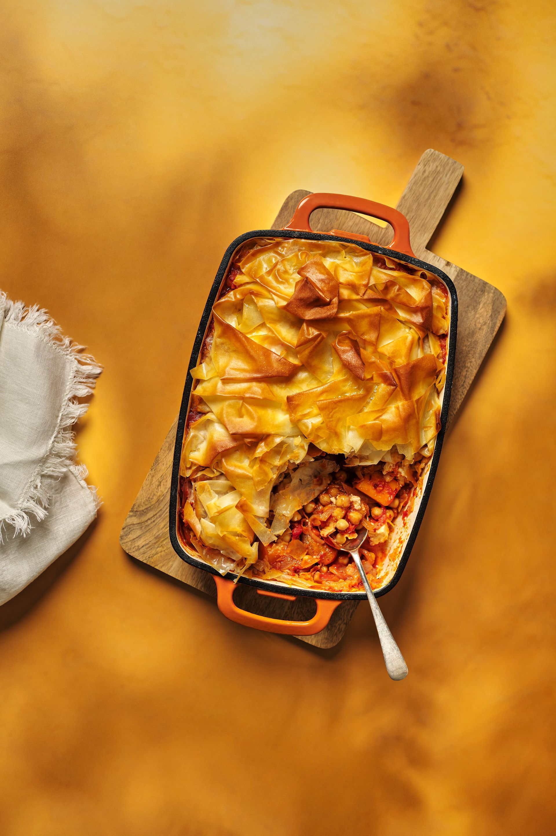A casserole dish with a spoon in it is on a wooden cutting board.