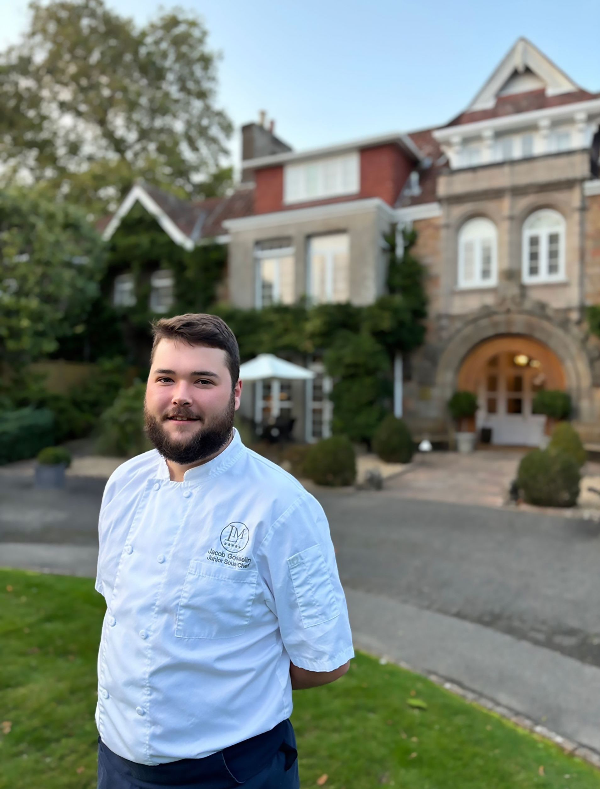 A man with a beard is standing in front of a large house.