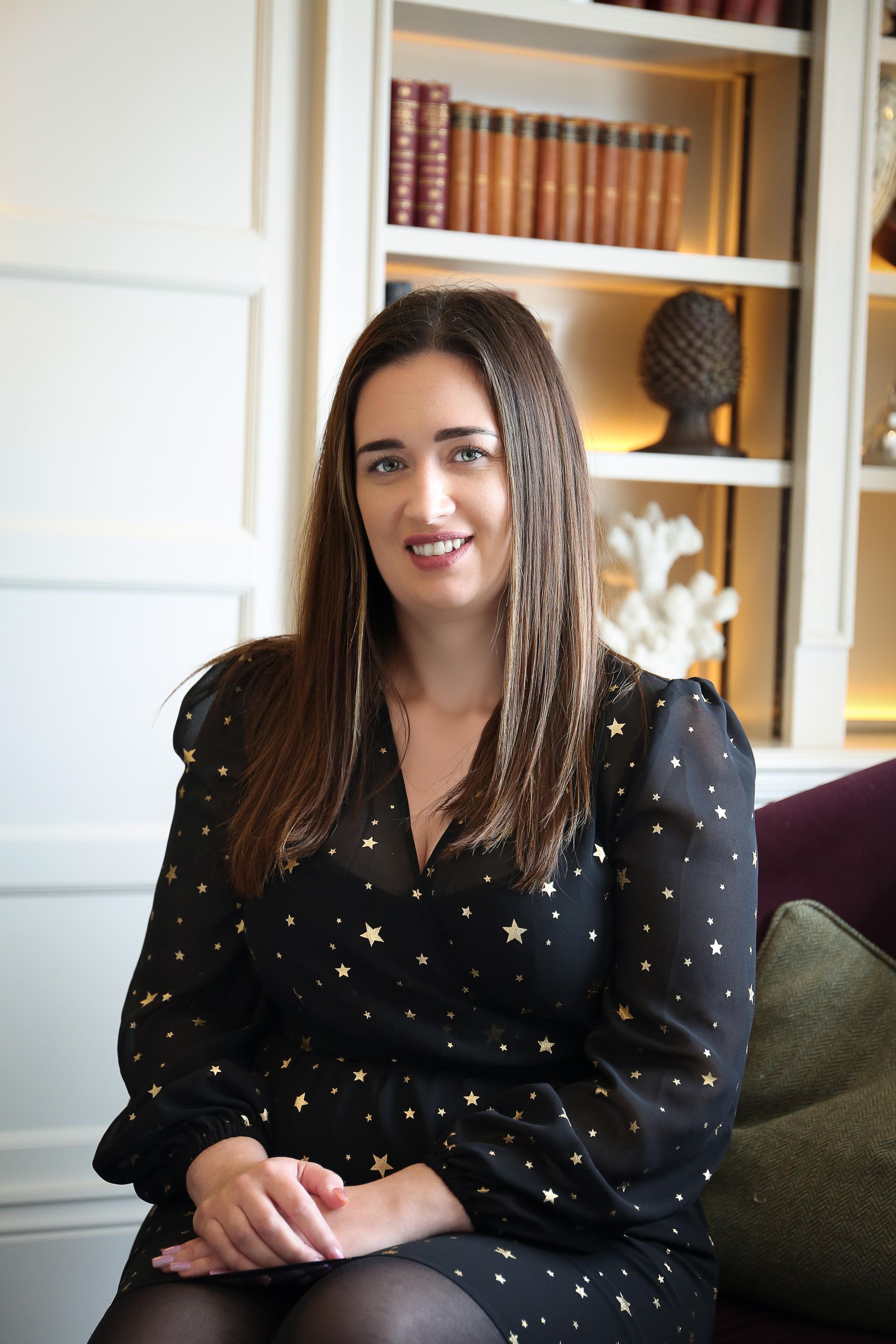 Woman with brown hair smiles, wearing a black dress with gold stars, sitting in a room with books and a sculpture.