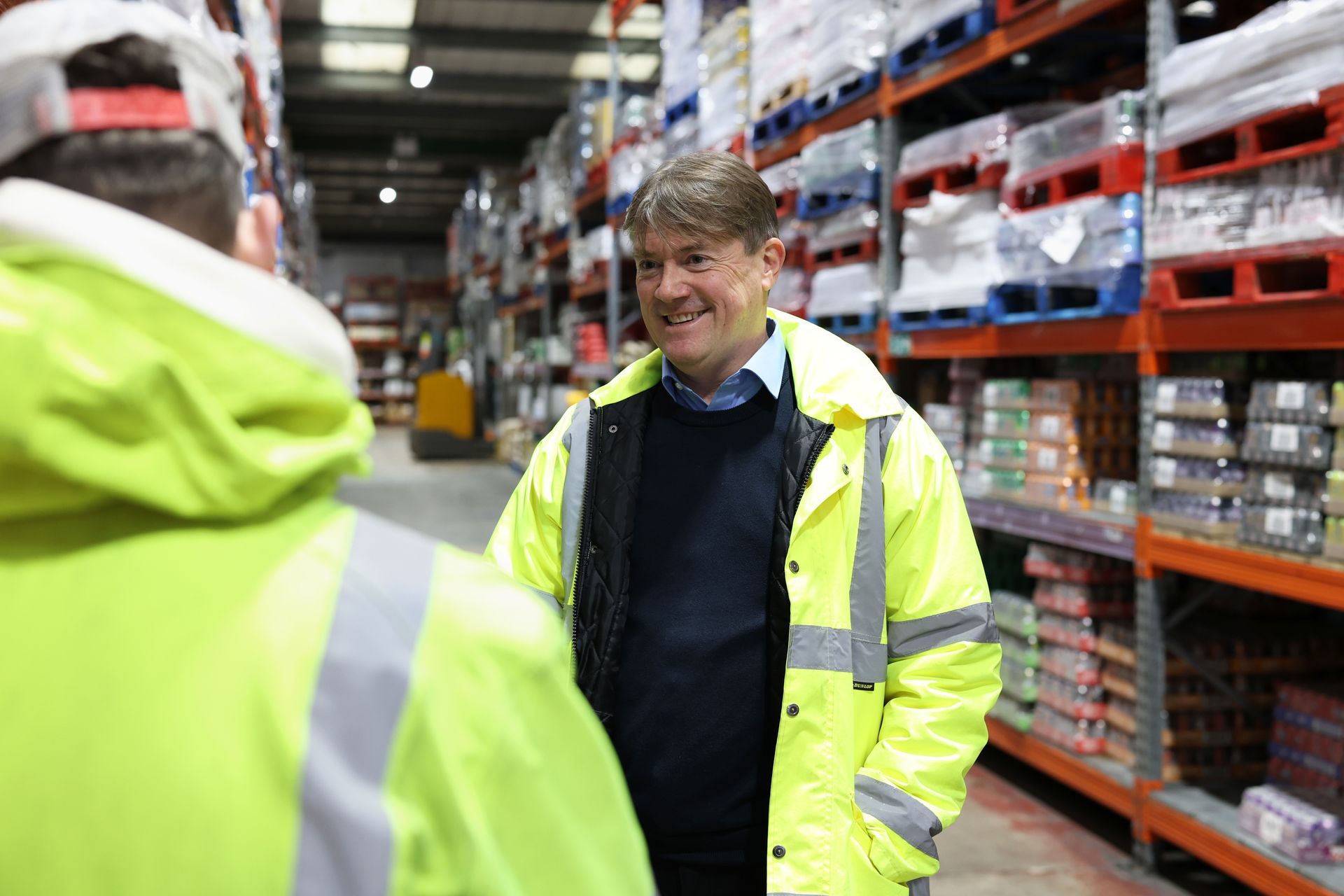A man in a yellow jacket is talking to another man in a warehouse.