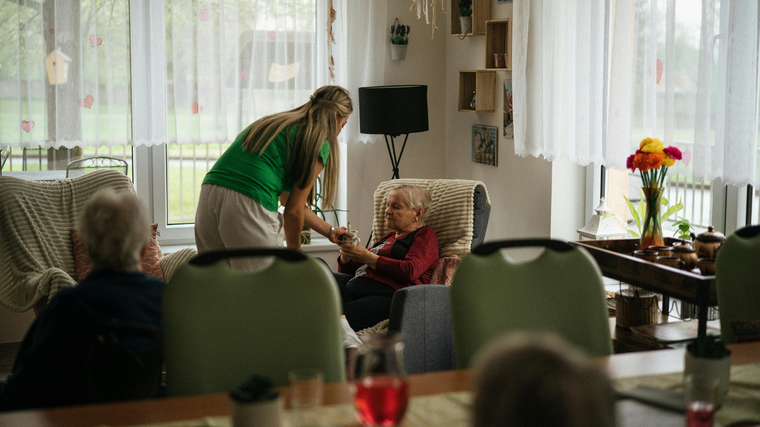 Caregiver leaning to talk with seated resident in bright living room, with flowers and large windows nearby