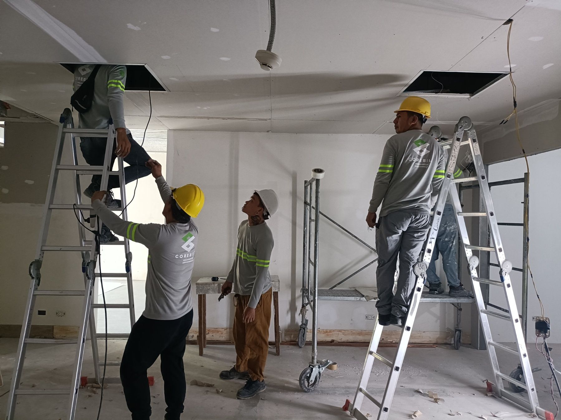 Construction workers installing electrical wiring in a white-walled room, using ladders to reach the ceiling.