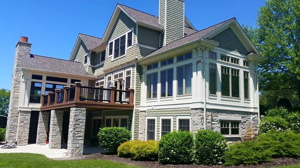 Large two-story house with light green siding, stone accents, and a deck, on a sunny day.