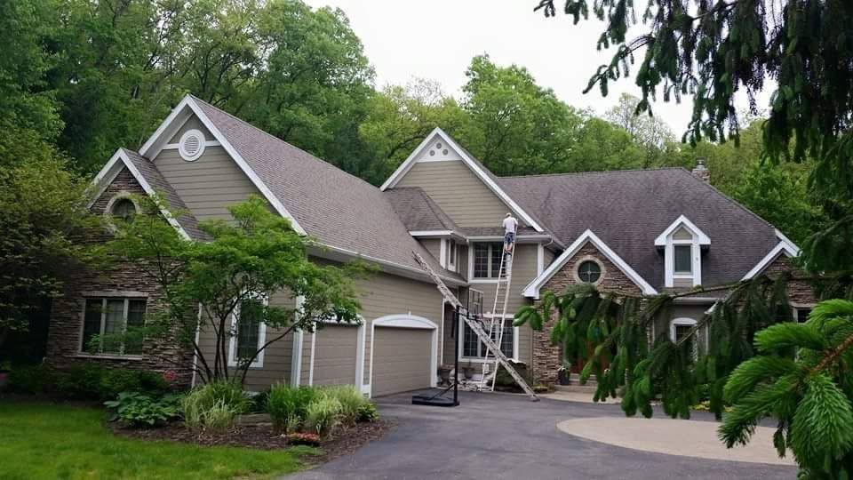 House with tan siding, stone accents, gray roof, and a circular driveway, surrounded by trees.