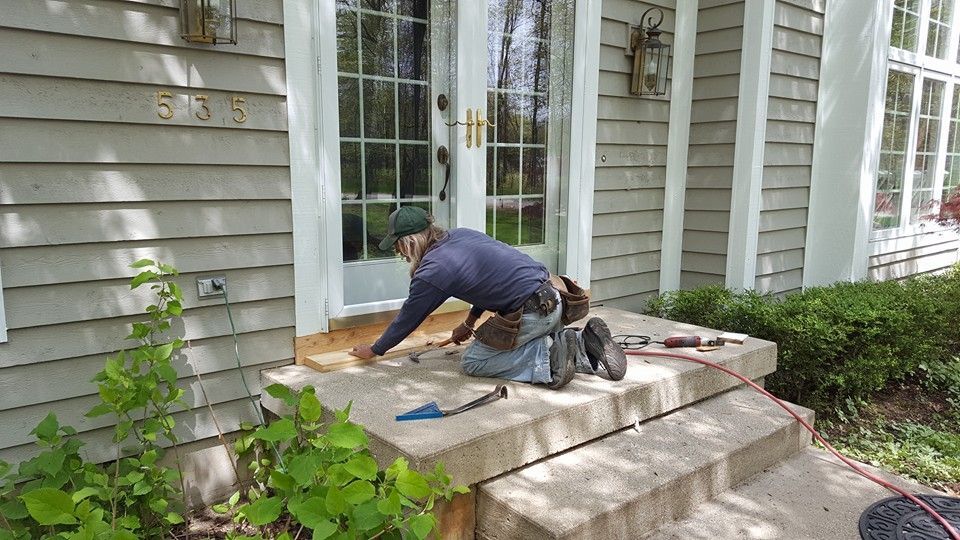 Man kneels on concrete steps outside a house, working on wood trim around a door.