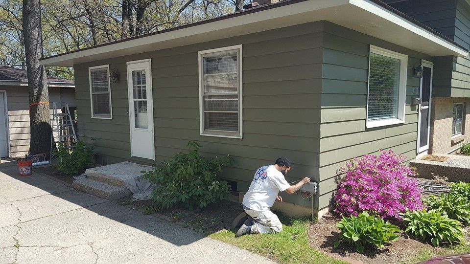 Person painting the exterior of a green house with white trim, kneeling on the grass.