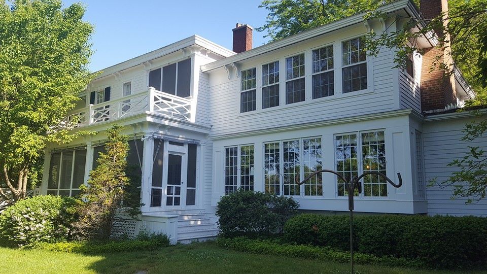 White house with screened porch, windows, and chimney on a sunny day; green lawn and trees.