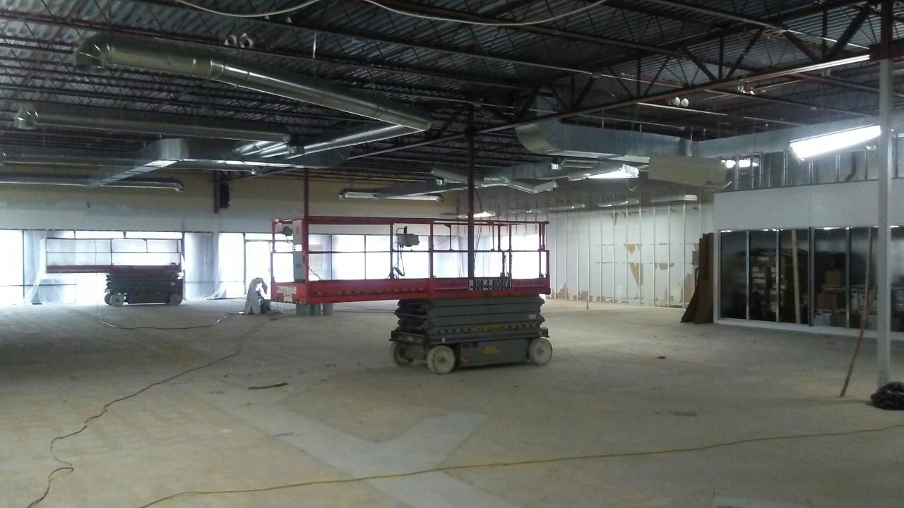 Interior construction site with lift and ductwork visible. Concrete floor, exposed ceiling, and white walls.