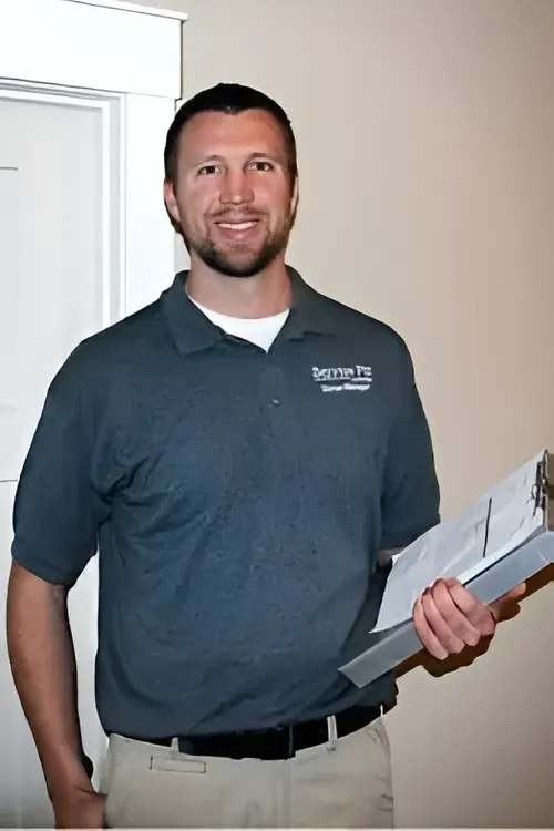 Man in gray polo shirt, tan pants, holding clipboard, smiling by a door.