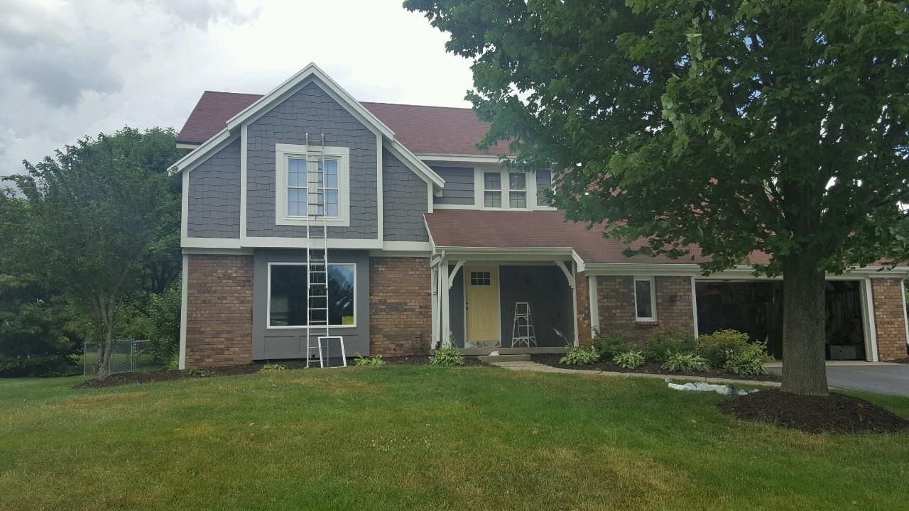 Two-story house, gray siding, red roof, brick base, ladder leaning against window, front yard with green grass.