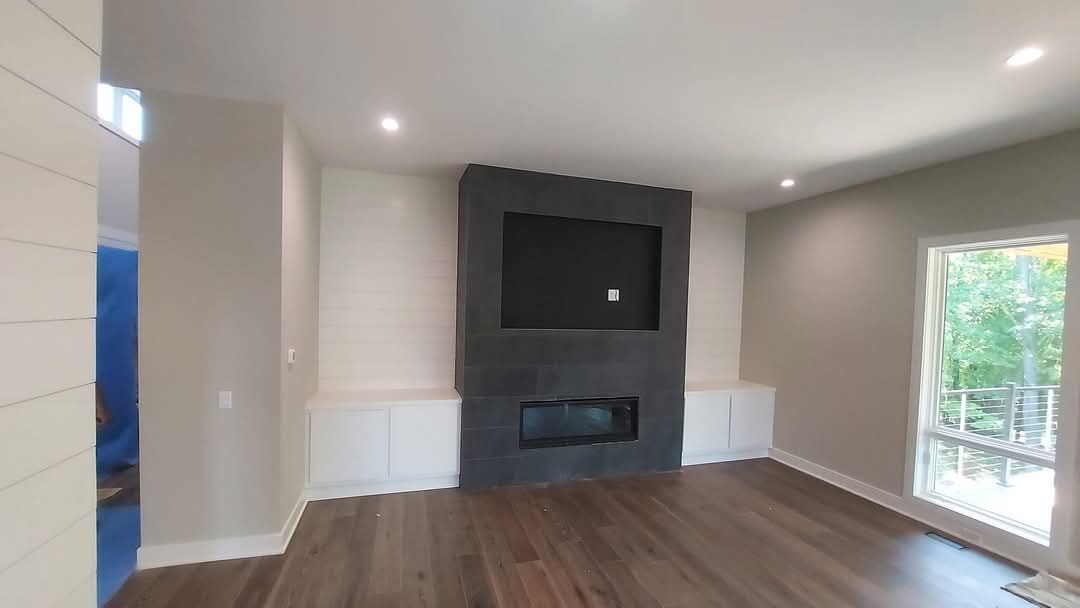 Living room with fireplace, mounted TV, built-in white cabinets, and wooden floors; neutral walls, and a large window.