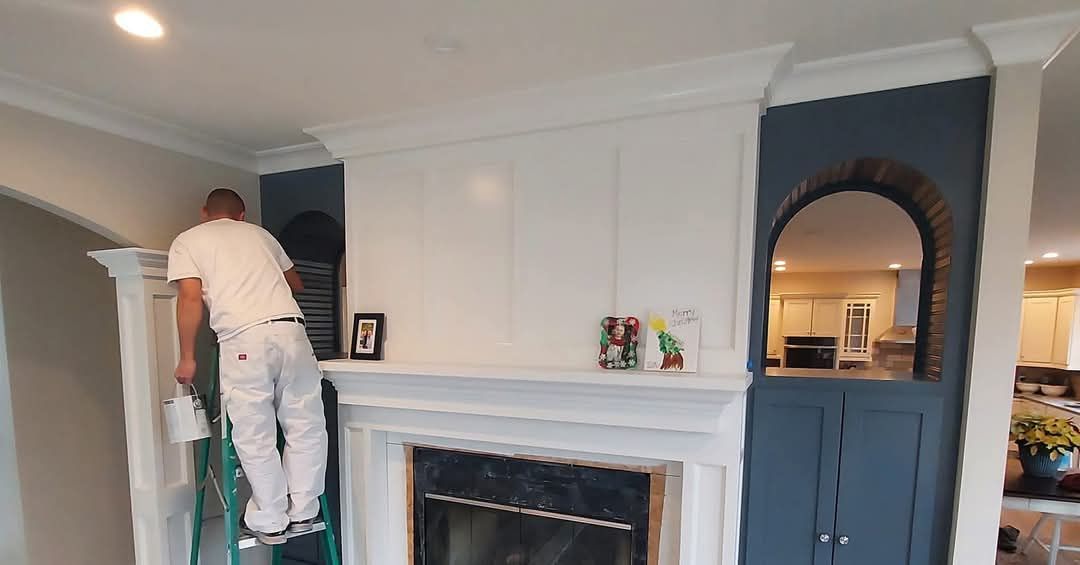 A person on a ladder paints a white fireplace and cabinet, with navy walls on either side.