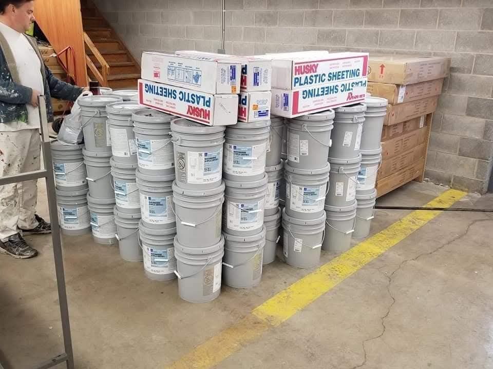 Person standing near a stack of gray paint buckets with boxes on top in a warehouse setting.