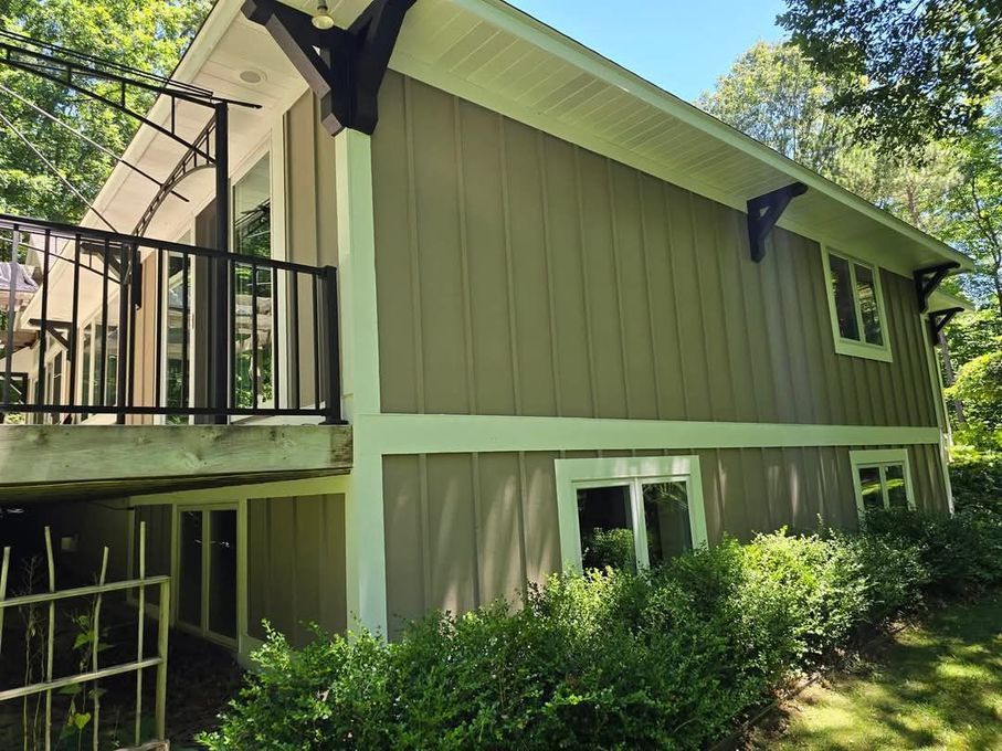 Two-story house with olive siding, black balcony, and white trim, surrounded by green foliage.