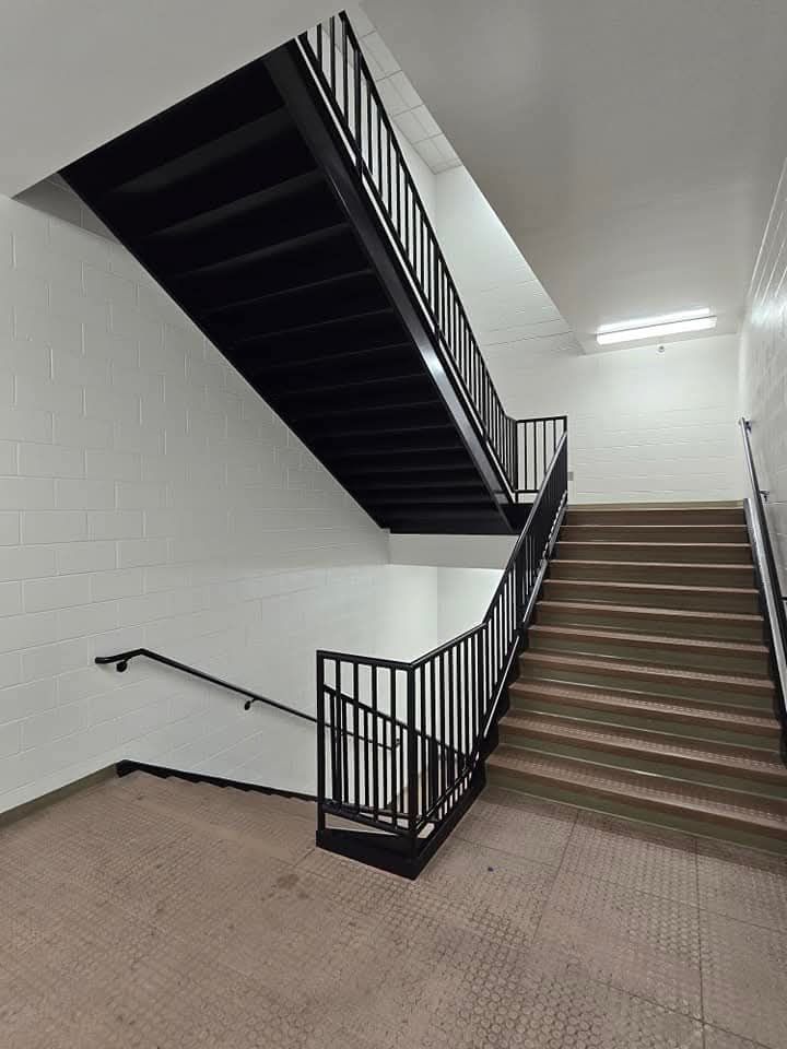 Stairwell with black metal handrails and steps, white tile walls, and tan flooring.