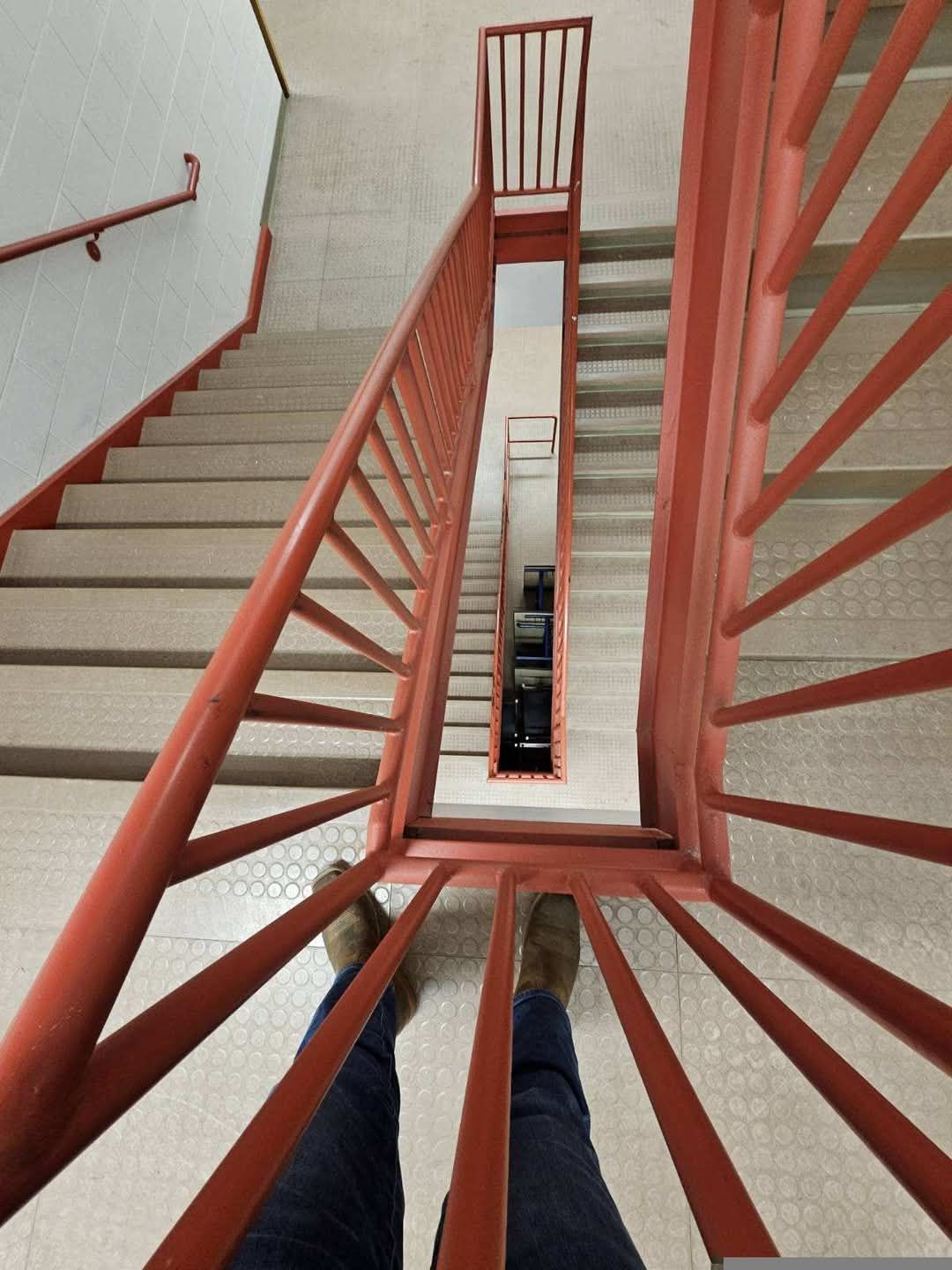 Looking down a red-railed stairwell. Person's feet in jeans and brown shoes are in the foreground.