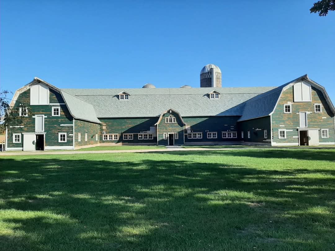 Green barn with a central silo, on a grassy field under a blue sky.