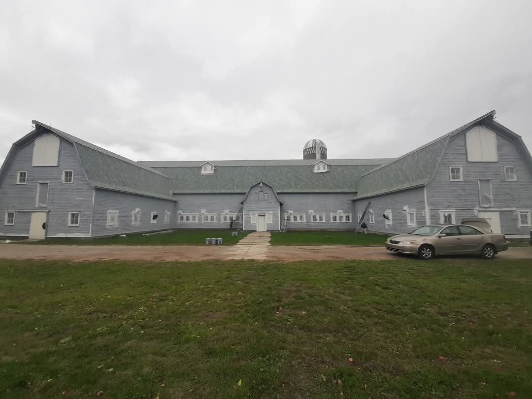 Gray barn with a central cupola, under an overcast sky. A car sits to the right.