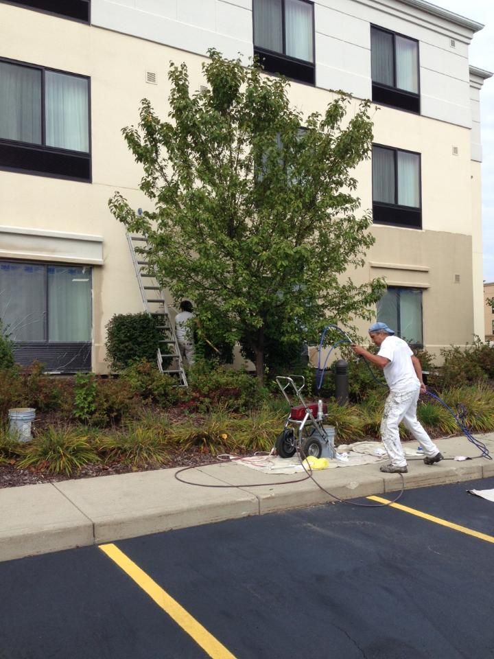 Painters work on a building exterior with a tree in between, using a paint sprayer.