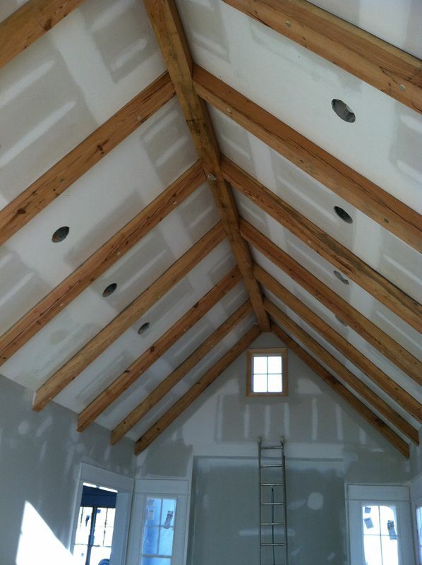 Interior view of a room under construction. Wooden beams form a vaulted ceiling, with drywall and recessed lighting.