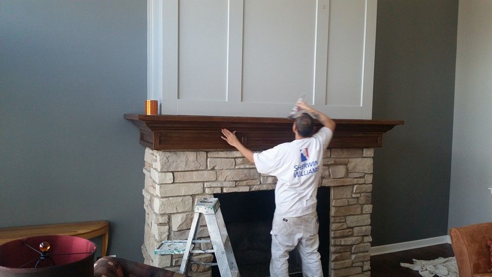 Man painting a fireplace mantel. Stone fireplace with white cabinet, gray walls.