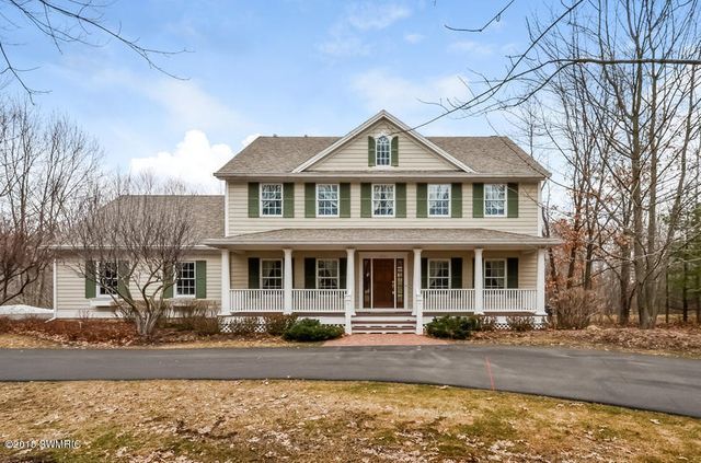 Two-story beige house with green shutters, a wrap-around porch, and a long driveway.
