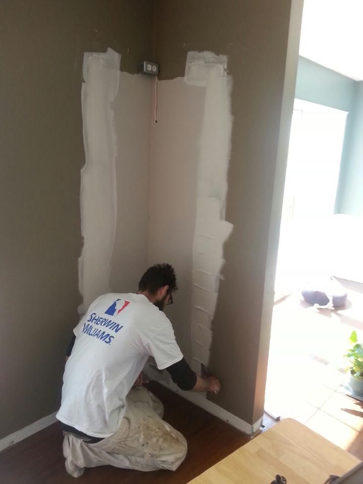 A painter in a white Sherwin-Williams t-shirt kneels, applying mud to a partially drywalled corner of a room.