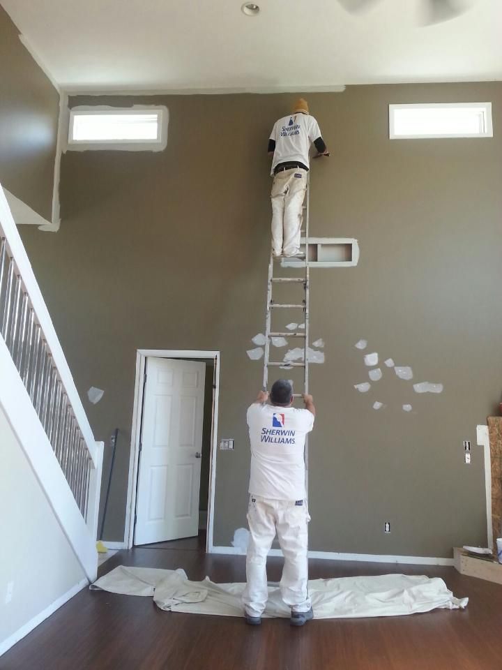 Two painters on a tall ladder painting a high brown wall; one is painting, other is supporting the ladder.