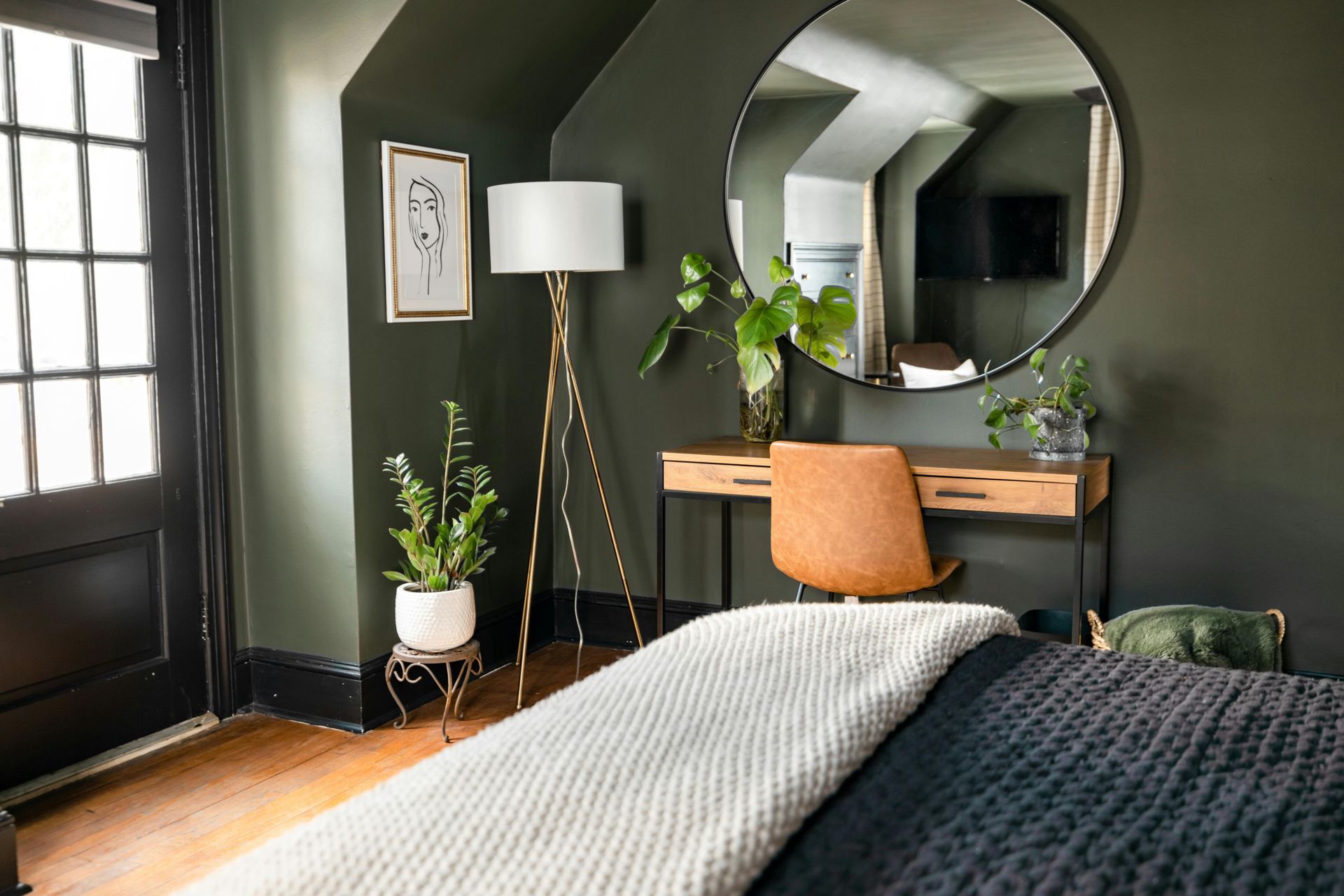 Bedroom with dark green walls, a gold desk and lamp, large round mirror, and a bed with a light throw blanket.