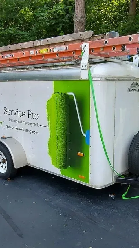 A white trailer with a Service Pro logo, ladder on top, green roller design, and parked on asphalt.