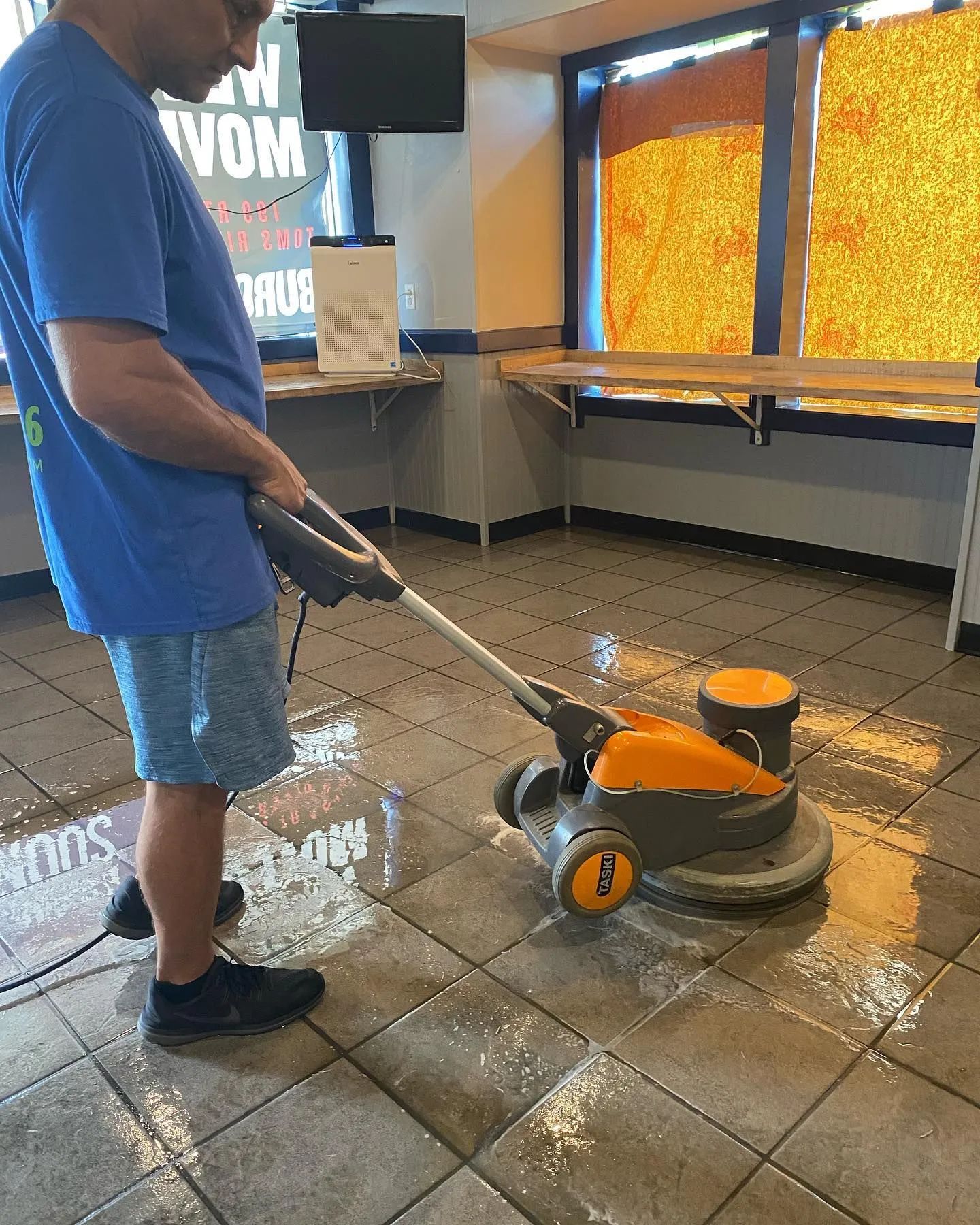 Man in blue shirt using floor cleaning machine on wet, tiled floor.