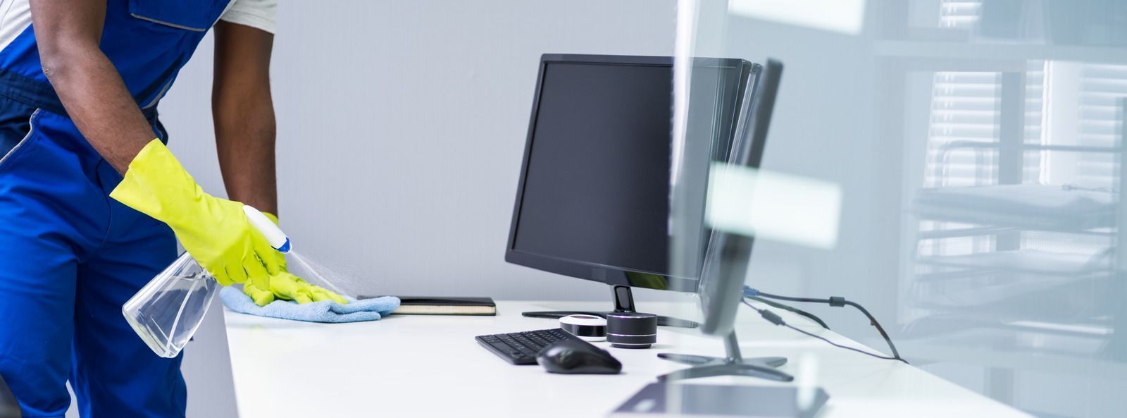 A person wearing yellow gloves cleaning a white office desk with a spray bottle and cloth.