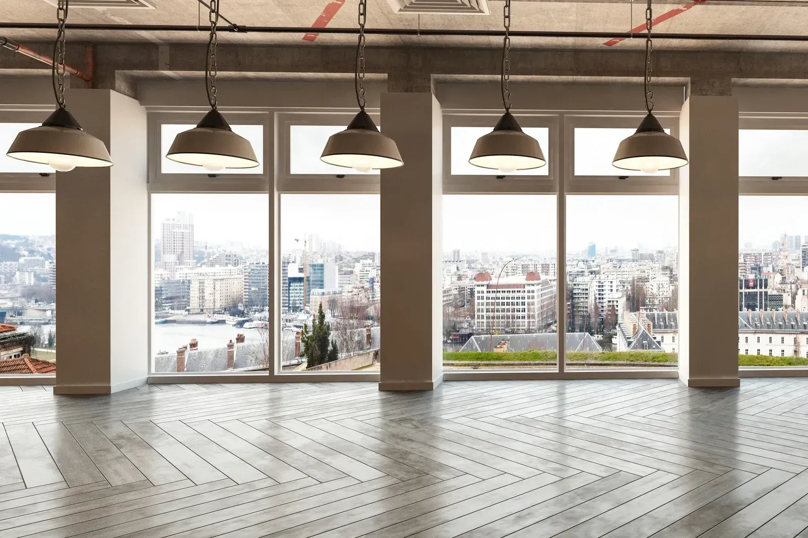 An empty room with large windows overlooking a city, overhead lights and light colored wooden floor.