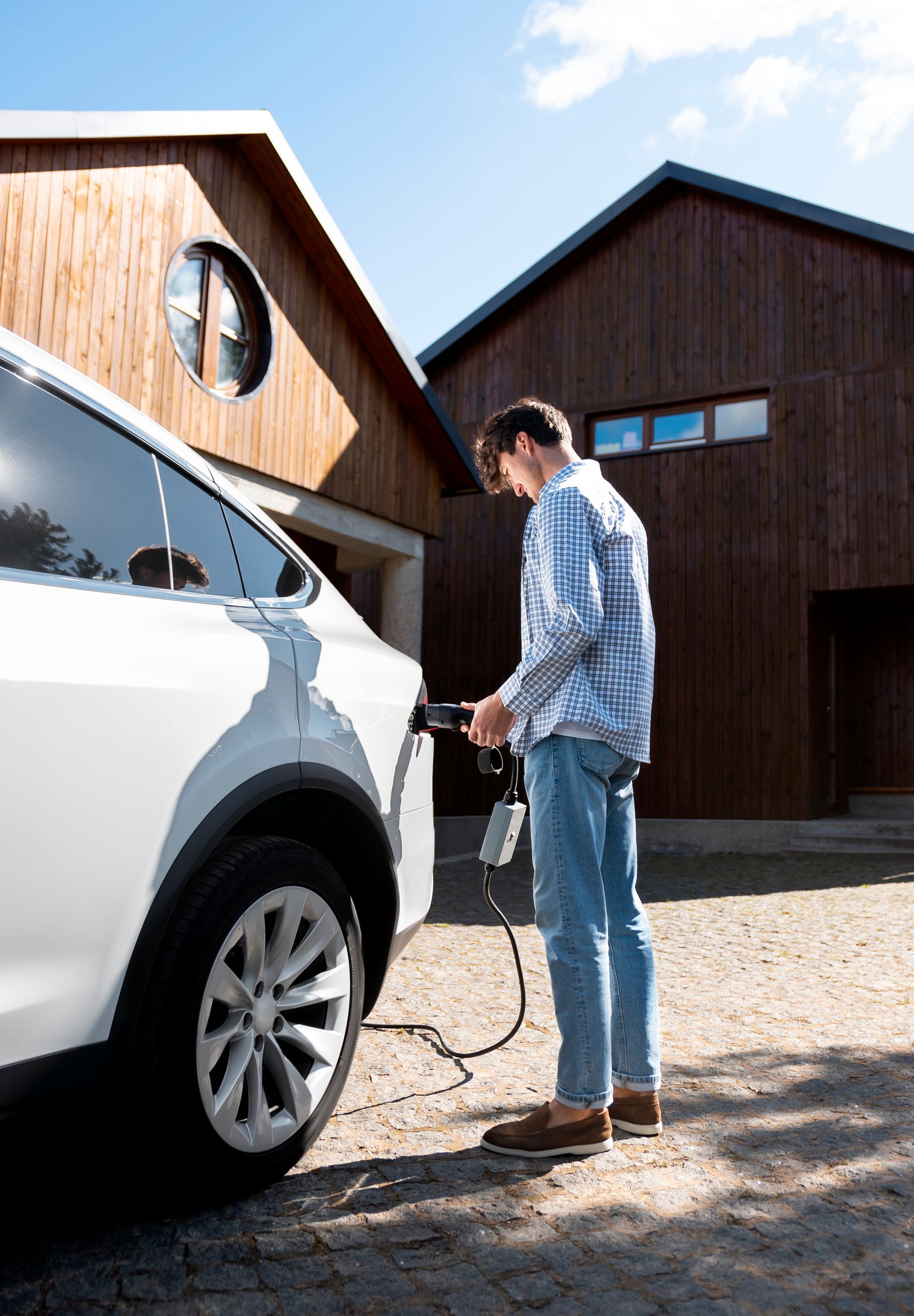 A man is charging his electric car in front of a wooden house.