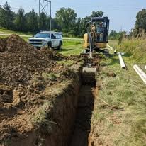 Backhoe digging a trench beside a roadside, with a white pickup and dirt piles nearby