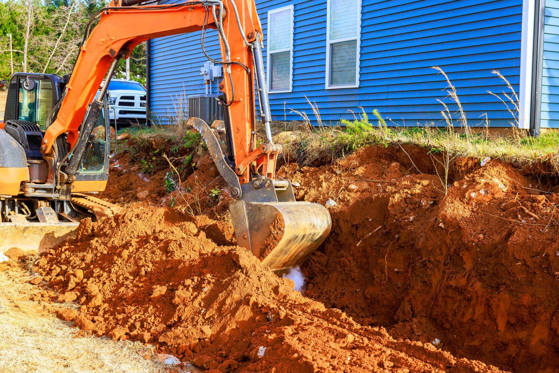 Orange excavator with front blade on a gravel path near a grassy riverbank and trees