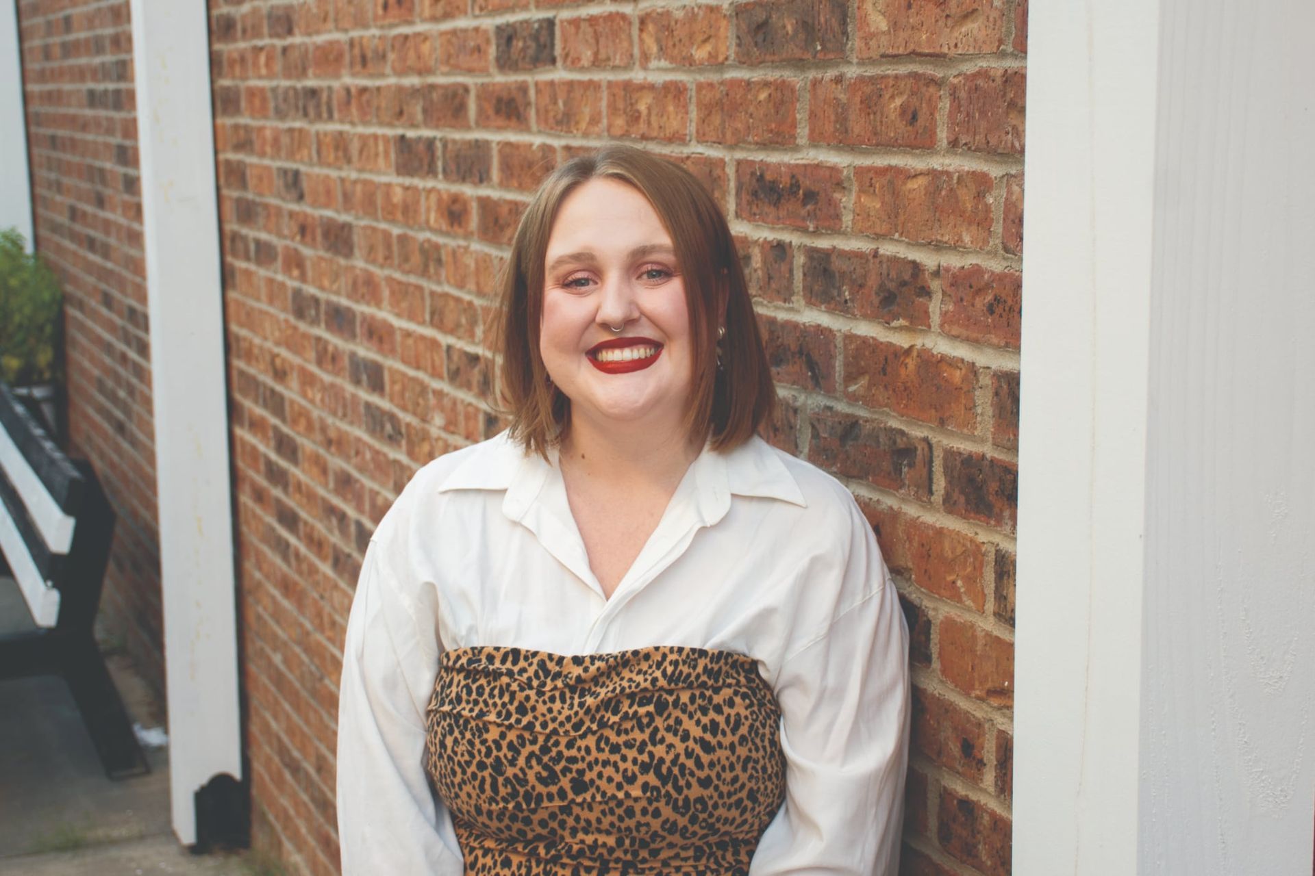 A smiling person with brown hair wears a white button-down shirt and leopard-print top, standing against a brick wall.