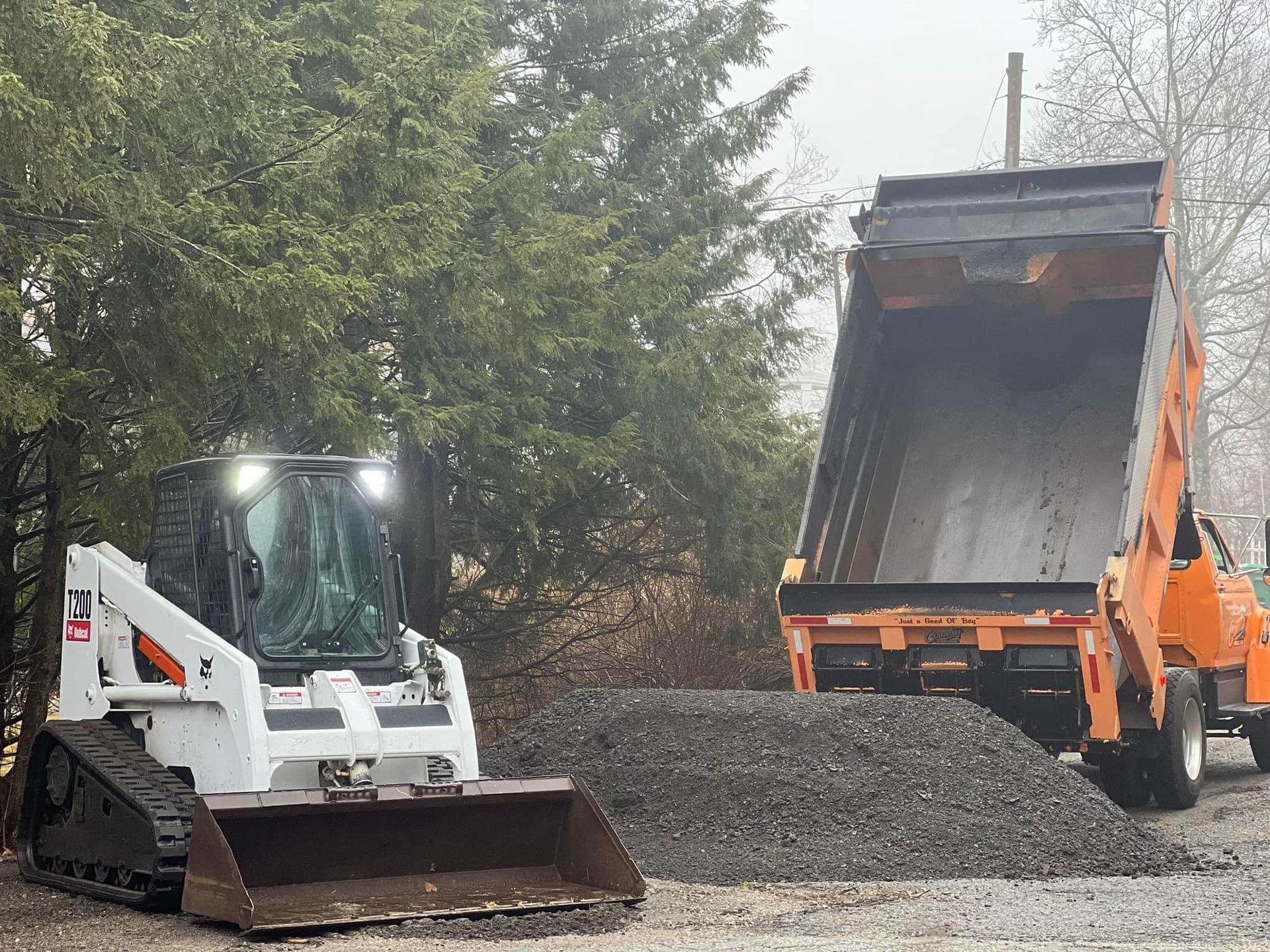 White Bobcat skid-steer loader next to orange dump truck and pile of gravel; outdoors.