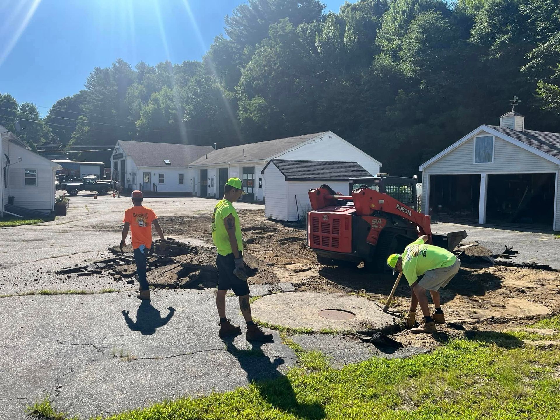 Construction workers removing asphalt with a skid steer, trees, and buildings in the background.
