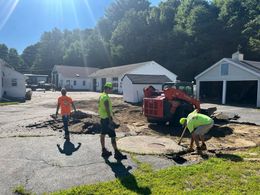 Construction workers removing asphalt; Kubota skid steer; buildings in the background; sunny day.