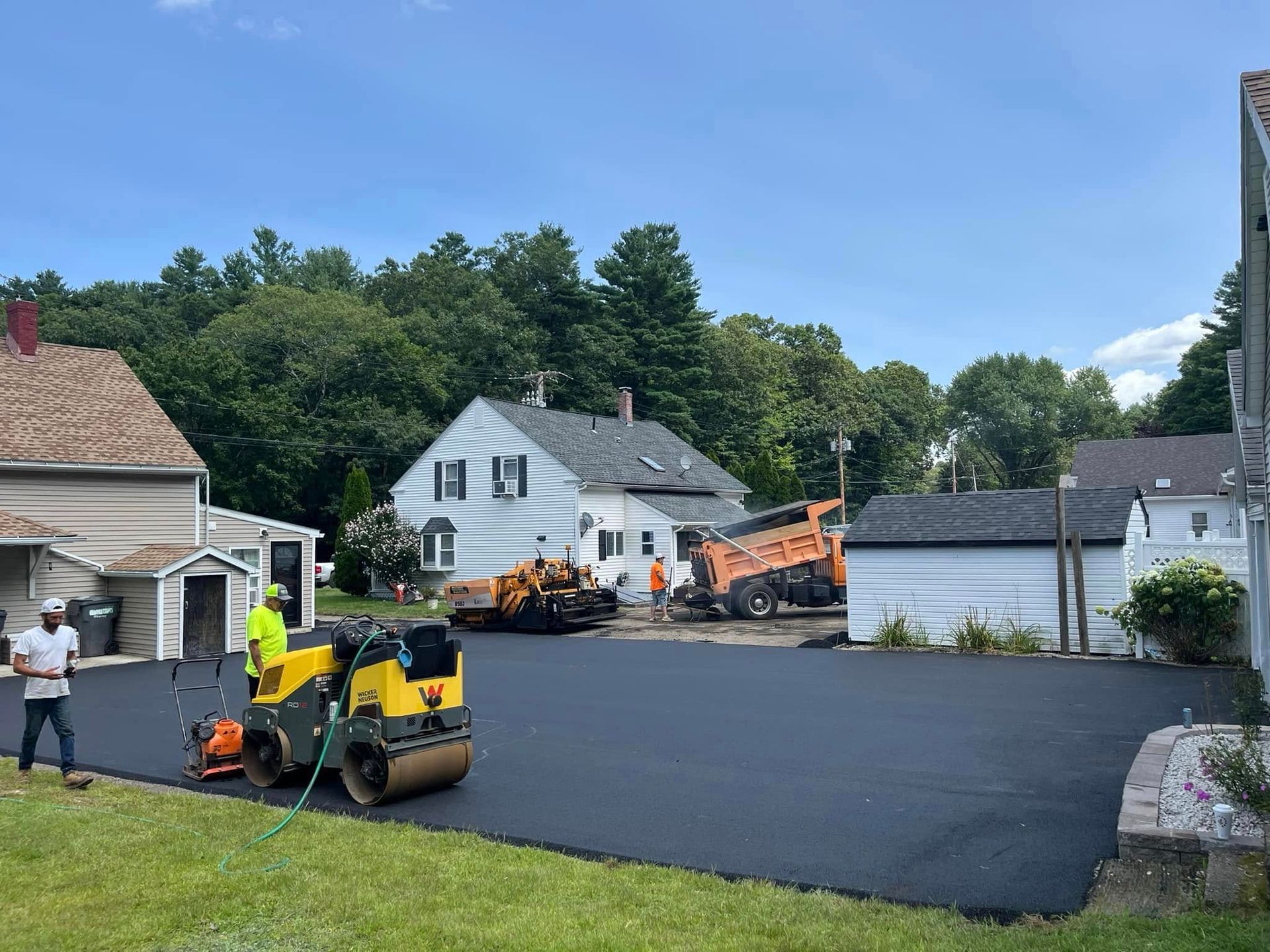 Asphalt paving in progress: workers, machinery on a residential driveway, houses in background, sunny day.
