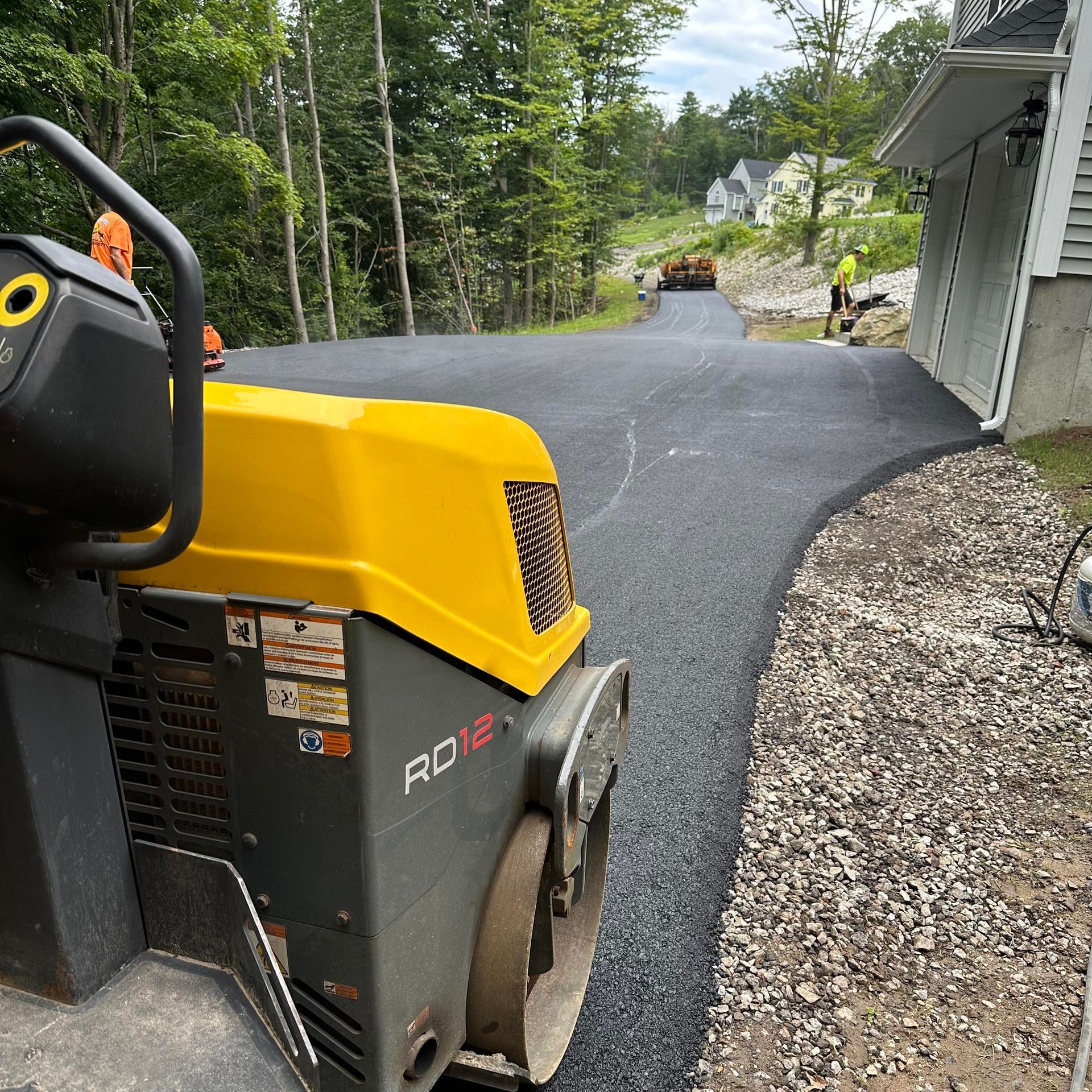 Asphalt paving a driveway, yellow roller in foreground, worker, trees in background.
