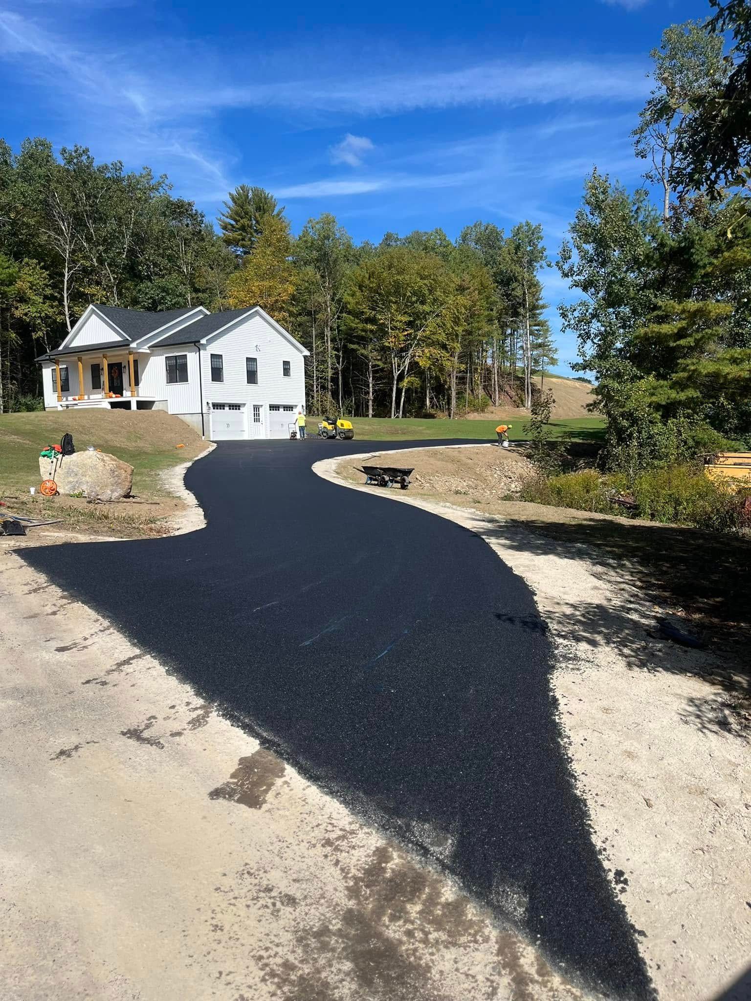 New asphalt driveway leading to a white house with a garage in a wooded area, blue sky.