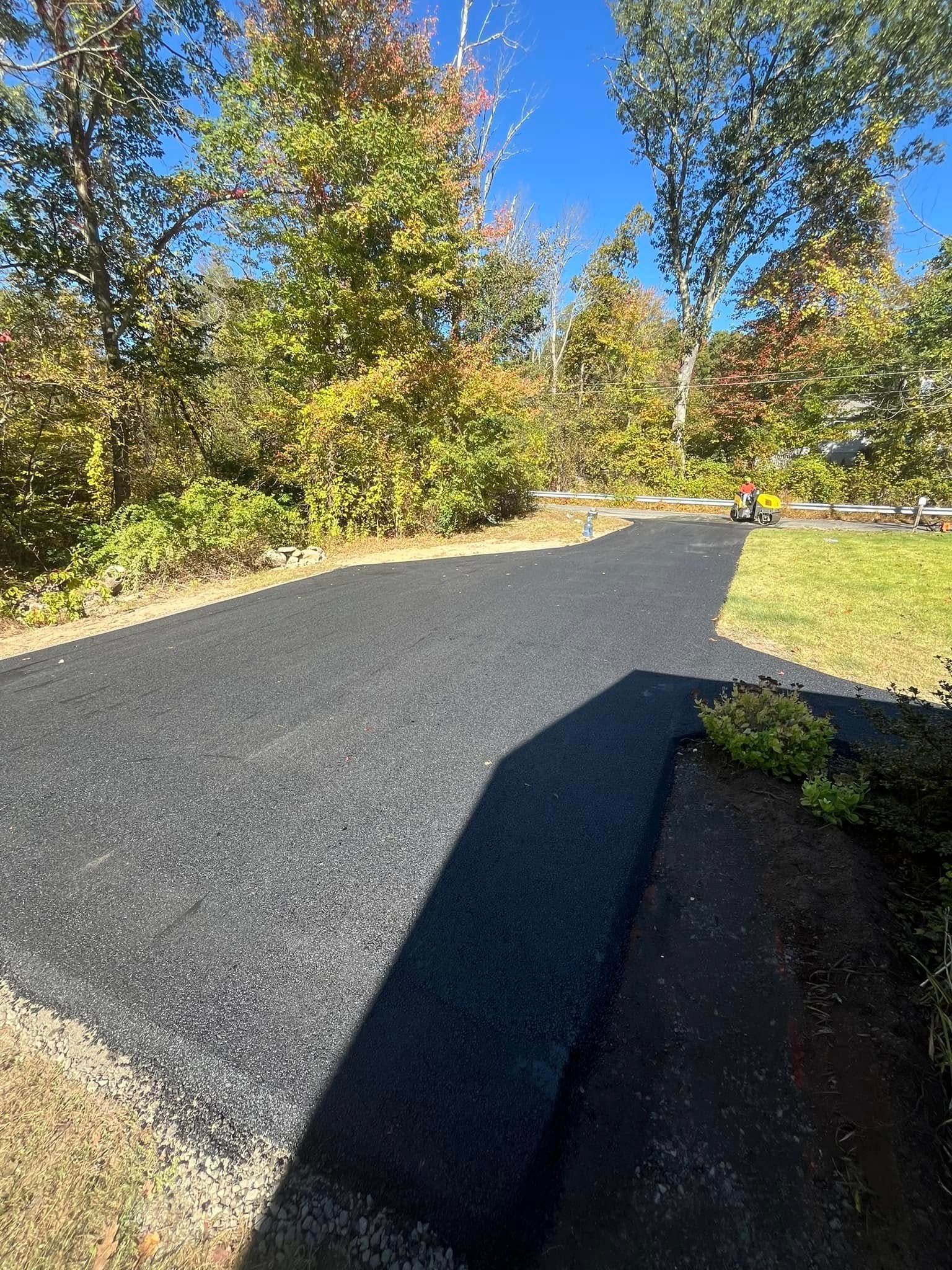 Black asphalt driveway surrounded by green grass and trees with some autumn foliage. Sunny day.