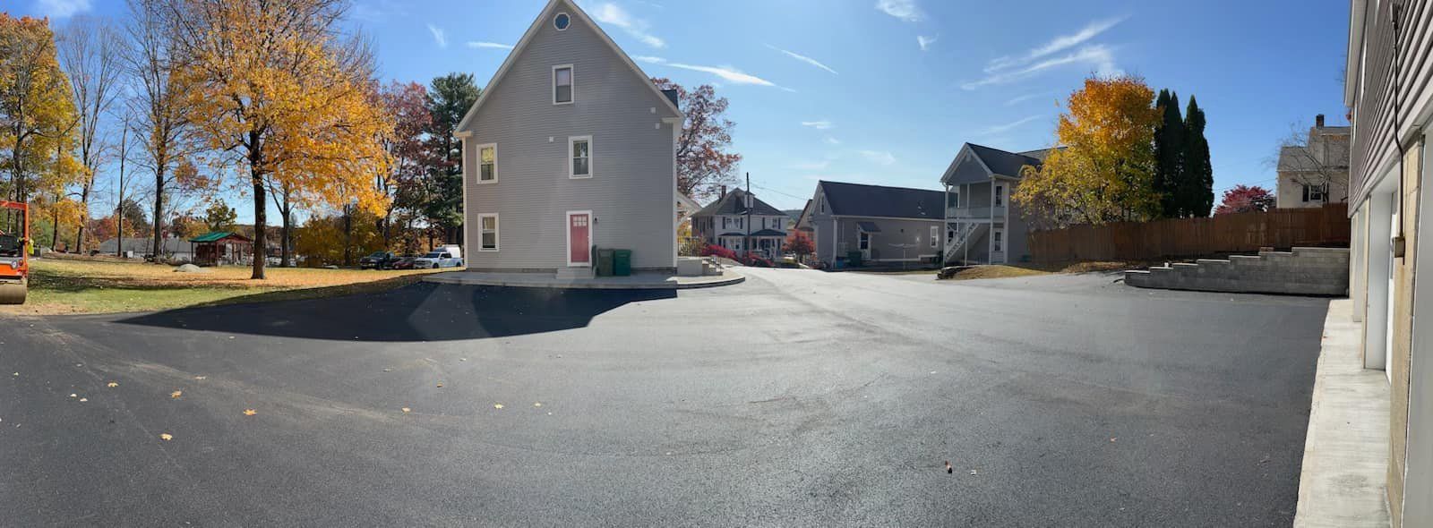 Paved lot with buildings on a sunny day; trees with yellow leaves in the background.